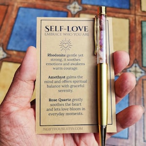 Op de afbeelding: Een goudkleurige pen met een doorzichtige huls gevuld met roze en paarse stenen wordt naast een kaart gehouden. De kaart leest "SELF-LOVE EMBRACE WHO YOU ARE" en vermeldt de eigenschappen van Rodoniet, Amethist en Rozenkwarts.