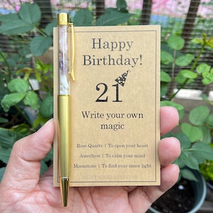 Op de afbeelding: Een gouden pen met een doorzichtige cilinder gevuld met kleine steentjes en een gouden clip, vastgehouden naast een kaart. De kaart leest "Happy Birthday! 21" en "Write your own magic". De kaart vermeldt ook de eigenschappen van rozenkwarts, amethist en maansteen.