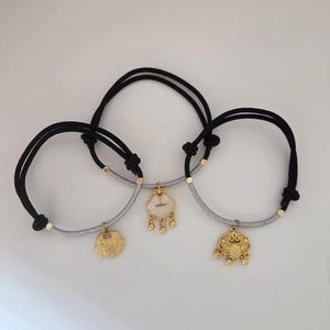 May include: Three bracelets with black cords and gold-colored charms. The charms have different shapes, including a cloud, a flower, and a decorative lock. The bracelets have a silver-gray band and gold accents.