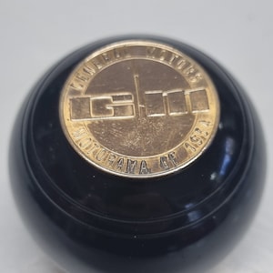 Puede incluir: Un pomo de plástico negro con un emblema de metal dorado que dice "General Motors Motorama of 1954".