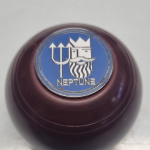 Könnte beinhalten: Ein dunkelbrauner Holz-Knopf mit einem blauen und silbernen Emblem. Das Emblem zeigt einen Dreizack und einen gekrönten Kopf mit Bart. Der Text "NEPTUNE" befindet sich unter dem Emblem.