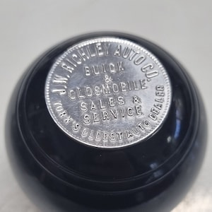 Könnte beinhalten: Ein schwarzer, runder Schaltknauf mit einem silbernen Emblem. Das Emblem lautet "J.W. Richley Auto Co. Buick & Oldsmobile Sales & Service, York's Oldest Auto Dealer."