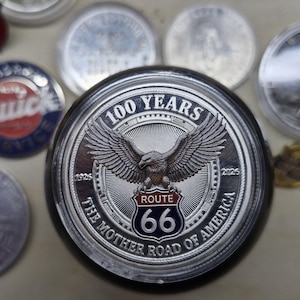 May include: A circular silver-toned commemorative coin featuring a central eagle with wings spread above a Route 66 shield emblem. The text reads 100 YEARS, ROUTE 66, and THE MOTHER ROAD OF AMERICA, with the years 1926 and 2026 flanking the central design.