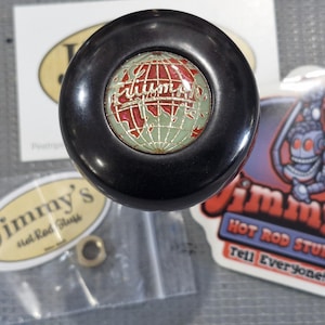 May include: A black Triumph shift knob with a red and green globe design. The knob is surrounded by stickers from Jimmy's Hot Rod Stuff and a small bag with hardware.