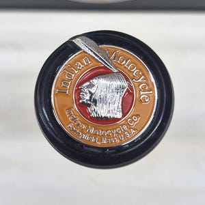 Puede incluir: Un pomo de palanca de cambios circular con el logotipo de Indian Motorcycle, que presenta un perfil plateado de una persona con tocado sobre fondo rojo, rodeado por el texto naranja Indian Motocycle Co. Springfield, Mass U.S.A.