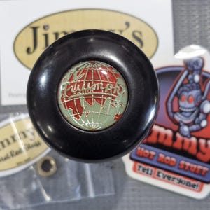 May include: A black Triumph shift knob with a red and green globe design. The word "Triumph" is written across the globe. The knob is surrounded by stickers that say "Jimmy's Hot Rod Stuff".