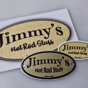 May include: Three oval-shaped metal signs with a gold background and black text. The signs read "Jimmy's Hot Rod Stuff".