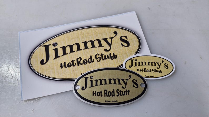 May include: Three oval-shaped metal signs with a gold background and black text. The signs read "Jimmy's Hot Rod Stuff".