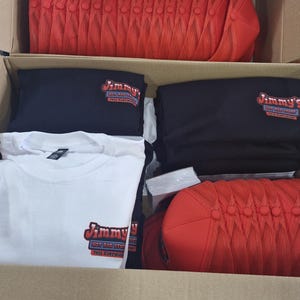 May include: A cardboard box filled with red mesh trucker hats, black t-shirts, and white t-shirts. The t-shirts have the logo "Jimmy's Hot Rod Stuff" printed on them.