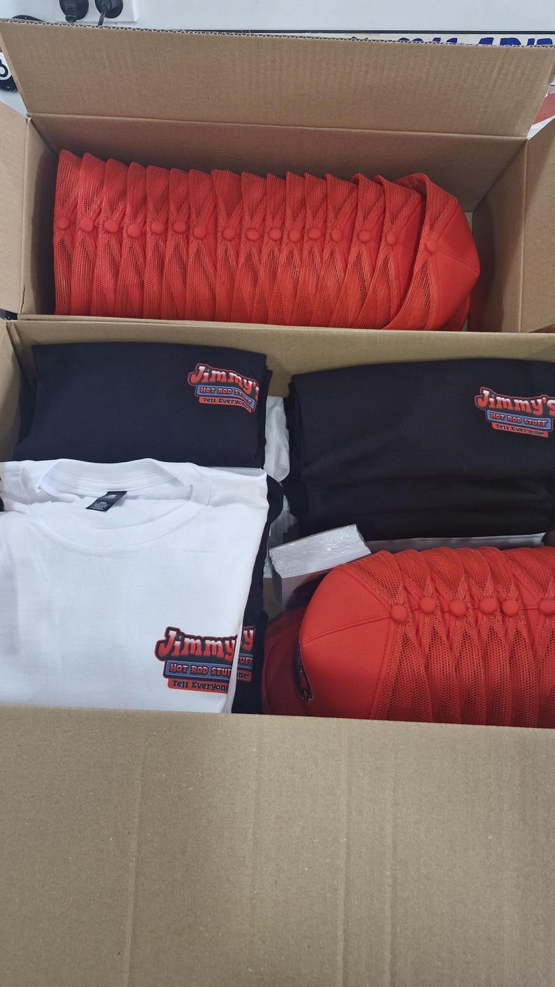 May include: A cardboard box filled with red mesh trucker hats, black t-shirts, and white t-shirts. The t-shirts have the logo "Jimmy's Hot Rod Stuff" printed on them.
