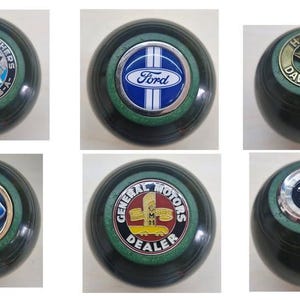 May include: Six dark green lawn bowls, each with a different automotive or motorcycle brand logo. Logos include Dodge Brothers, Ford, Harley-Davidson, General Motors, and others. Each bowl has a diameter of approximately 12 cm.