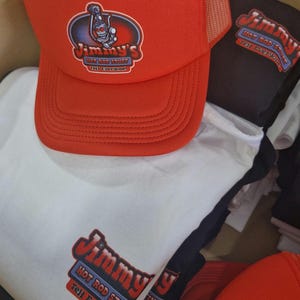 May include: Red and black trucker hat with a white logo that says "Jimmy's Hot Rod Stuff Tell Everyone!" The hat is on top of a white t-shirt with the same logo.