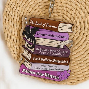 May include: A clear acrylic keychain depicting a stack of fantasy books with titles such as "The Book of Brennan" and "Dragon Rider's Codex." A black dragon silhouette is on the books. The keychain has a silver metal clasp.