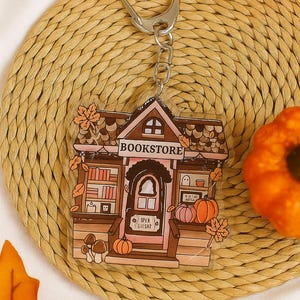 May include: A bookstore-themed keyring with a silver clasp. The acrylic charm features a brown and pink building with the word "BOOKSTORE" above the door. Pumpkins, mushrooms, and autumn leaves decorate the design. The keyring is on a woven mat.