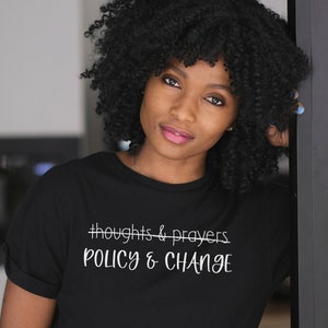 May include: A black t-shirt with white text that reads "#thoughts & prayers" with a line through it and "POLICY & CHANGE" underneath.