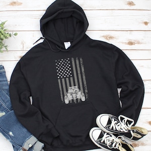 Puede incluir: Sudadera con capucha negra con un diseño de bandera estadounidense desgastado que presenta una silueta gris de un vehículo todoterreno.