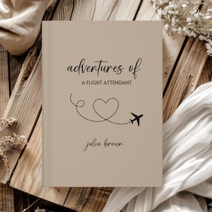 May include: A beige book with the title "adventures of a flight attendant" in black script. A dotted line forms a heart shape with a small airplane. The author's name, "julia brown," is below. The book is on a wooden surface.