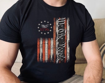 May include: Black t-shirt with a distressed American flag design and the text "We the People" printed vertically on the right side of the flag. The flag has a circle with 13 stars and the year "1776" printed inside.