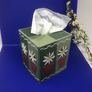 May include: A green and white tissue box cover with a floral design. The cover features three potted flowers with white petals and yellow centres. The tissue box cover is made of fabric and has a square shape.