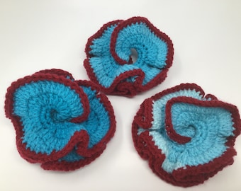 May include: Three crocheted flower-shaped ornaments in blue and burgundy. The ornaments are made with a textured yarn and have a ruffled edge.