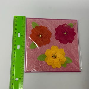 May include: A pink greeting card with glitter and three paper flowers. The flowers are orange, pink, and yellow. The card says "Papyrus" in the top right corner.