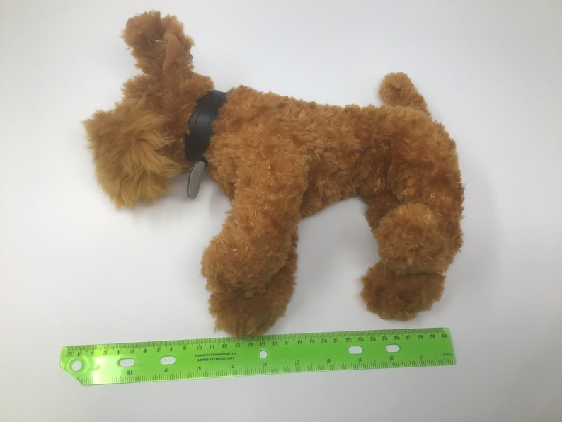 May include: A brown plush toy dog with a black leather collar and a tag. The dog is lying on its side with its head turned to the left. The dog is about 12 inches long.