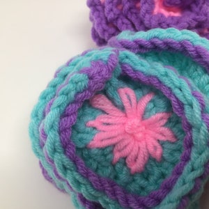 K&ouml;nnte beinhalten: Nahaufnahme einer geh&auml;kelten Blume mit pinkfarbenem Zentrum und t&uuml;rkisfarbenen und violetten Bl&uuml;tenbl&auml;ttern.