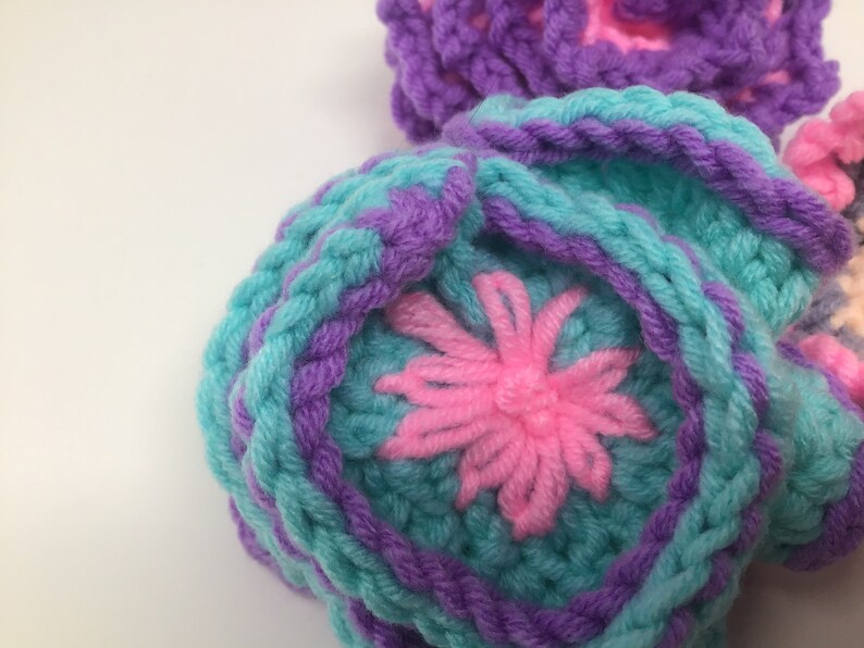 K&ouml;nnte beinhalten: Nahaufnahme einer geh&auml;kelten Blume mit pinkfarbenem Zentrum und t&uuml;rkisfarbenen und violetten Bl&uuml;tenbl&auml;ttern.