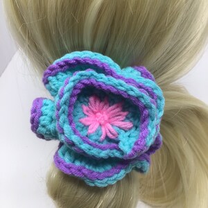 K&ouml;nnte beinhalten: Ein geh&auml;keltes Haargummi in T&uuml;rkis und Lila mit einer pinkfarbenen Blume in der Mitte.