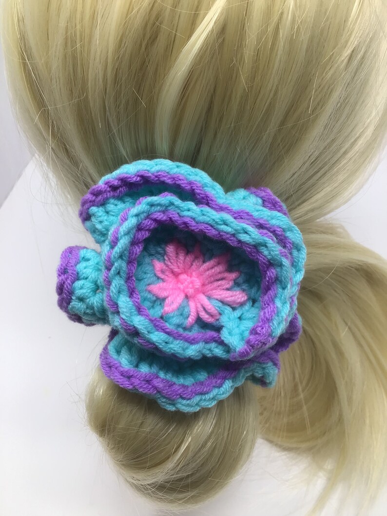 K&ouml;nnte beinhalten: Ein geh&auml;keltes Haargummi in T&uuml;rkis und Lila mit einer pinkfarbenen Blume in der Mitte.