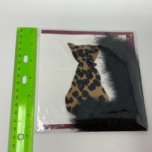 May include: A decorative cat-shaped item with a leopard print design, black feathers, and a purple accent. The item is packaged in clear plastic and includes the text "PAPYRUS". A green ruler is placed next to the item.