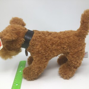 May include: A brown plush toy dog with a black leather collar and a red tag that says "GUND" on it. The dog is standing on a white surface.