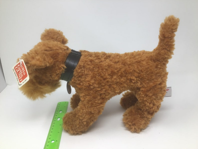 May include: A brown plush toy dog with a black leather collar and a red tag that says "GUND" on it. The dog is standing on a white surface.
