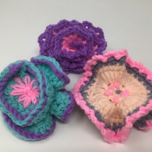 K&ouml;nnte beinhalten: Drei geh&auml;kelte Blumen in Rosa, Lila und T&uuml;rkis mit einem grauen und rosafarbenen Rand. Die Blumen sind mit verschiedenen H&auml;kelmustern gearbeitet und haben einen 3D-Effekt.