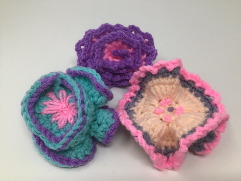 K&ouml;nnte beinhalten: Drei geh&auml;kelte Blumen in Rosa, Lila und T&uuml;rkis mit einem grauen und rosafarbenen Rand. Die Blumen sind mit verschiedenen H&auml;kelmustern gearbeitet und haben einen 3D-Effekt.