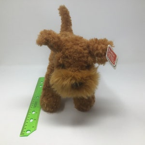 May include: A brown plush toy dog with a shaggy coat. The dog has a small black nose and brown eyes. The toy has a red and white tag that says "Gotta Getta Gund".