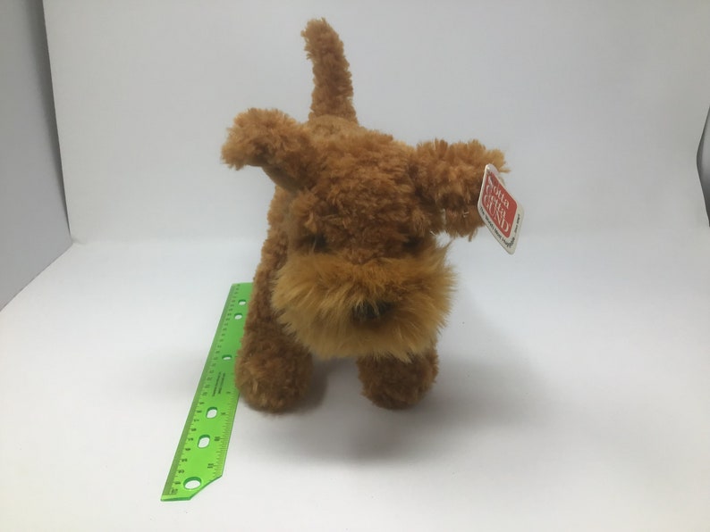 May include: A brown plush toy dog with a shaggy coat. The dog has a small black nose and brown eyes. The toy has a red and white tag that says "Gotta Getta Gund".