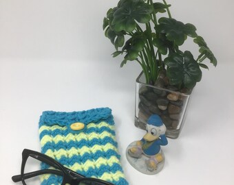 May include: A crocheted teal and yellow glasses case with a yellow button. A pair of black glasses is inside the case. A small figurine of a cartoon duck is in the background.