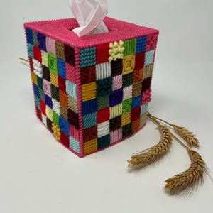 May include: A square tissue box cover in a patchwork design. The box is primarily pink with a top opening. The patchwork is made of various colors including red, blue, green, yellow, and white. Wheat stalks are on the side.