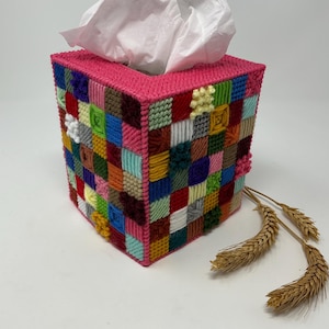 May include: A handmade tissue box cover with a patchwork design. The box is primarily pink with a square opening at the top. The sides are covered in a variety of colorful squares. Wheat stalks are placed near the box.