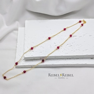 May include: A gold chain necklace with small, round, red beads. The necklace is on a white surface. Rebel Rebel Jewellery.