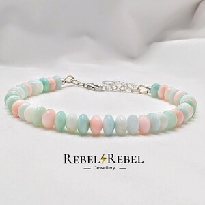 May include: A beaded bracelet featuring pastel-colored beads in shades of pink, blue, and white. The bracelet is strung on a silver chain with a clasp. The brand name "REBEL REBEL Jewellery" is printed below the bracelet.