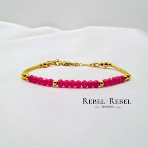 Puede incluir: Un brazalete de oro con una cadena de cuentas de piedras preciosas rosas. El brazalete tiene un diseño de cadena delicado y está acentuado con pequeñas cuentas doradas.