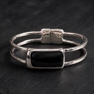 May include: A silver-toned cuff bracelet featuring a double-band design and a central rectangular black stone setting. The metal surface has a textured, hammered finish, and the piece includes a hinged clasp mechanism at the back of the band.