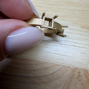 May include: A gold-colored ring setting with a matte, textured finish. The ring features a square-shaped central mounting with four prongs designed to hold a gemstone, along with additional small prongs extending from the sides of the band.