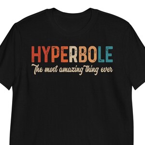 Puede incluir: Camiseta negra con la palabra "HYPERBOLE" en letras naranjas, blancas y turquesas. Debajo está la frase "The most amazing thing ever" en cursiva. La camiseta está hecha de un material suave.