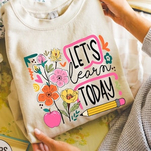 May include: A beige sweatshirt with the text "LET'S learn TODAY" in a pink and black design, accompanied by colorful floral and apple graphics. A yellow pencil graphic is also included. The sweatshirt is folded and held by a person.