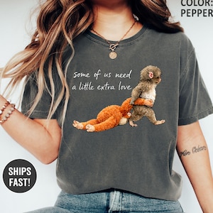 May include: Pepper-colored t-shirt featuring a graphic of a brown stuffed animal hugging a gray animal, with the text "some of us need a little extra love." The shirt is a casual style.