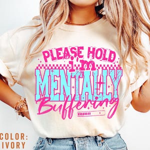 May include: Ivory-colored t-shirt with the text "PLEASE HOLD I'M MENTALLY BUFFERING" in pink and blue. The shirt features a retro design with a loading bar graphic. The color of the shirt is ivory.