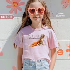 May include: A pale pink t-shirt featuring the phrase "some of us need a little extra love" and a graphic of a monkey hugging a stuffed toy. The shirt also has the text "SHIPS FAST!" and "COLOR: BLOSSOM".